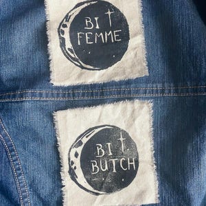 May include: A blue denim jacket with two white fabric patches. Each patch has a dark circle with a crescent moon and a cross, with the words "BI FEMME" and "BI BUTCH" printed in black.