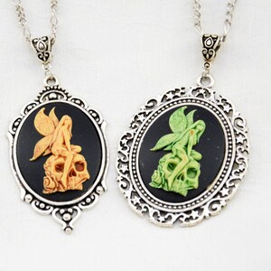 Könnte beinhalten: Zwei silberne Halsketten mit verzierten Rahmen und schwarzen Kameenanhängern. Jeder Anhänger zeigt eine weiße Fee, die auf einem Schädel sitzt, eine mit einer grünen Fee und die andere mit einer orangefarbenen Fee.