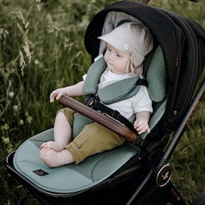 May include: A black stroller with a sage green seat liner and a brown leather-look bumper bar. A child sits inside wearing a white shirt, mustard-colored pants, and a beige sun hat. The stroller features a black canopy and a black frame.