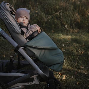 May include: A Joolz Geo stroller featuring a light gray frame and seat, equipped with a dark sage green footmuff. The stroller has black wheels and a structured design, providing a secure seating area for a child wearing a brown beanie and sweater.