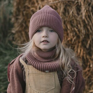 Peut inclure: Un enfant portant un bonnet en tricot côtelé mauve et une écharpe à col roulé assortie. L'enfant porte également une veste mauve et une salopette en velours côtelé beige. L'arrière-plan présente une botte de foin floue.