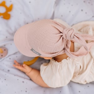 May include: A pink muslin baby hat with a tie at the top. The hat has a small label with the word "Bababie" on it.
