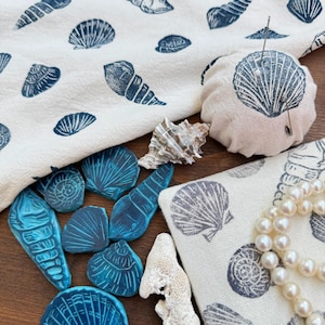 Puede incluir: Una variedad de artículos con temática de conchas marinas. Se muestra una tela blanca con estampados de conchas marinas azules, un alfiletero con diseño de concha marina y varios objetos azules con forma de concha marina. También hay un collar de perlas y un trozo de coral.