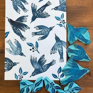 Può includere: Una stampa con un motivo di uccelli blu stilizzati e foglie su sfondo bianco. Diversi timbri di uccelli e foglie blu sono disposti attorno alla stampa su una superficie di legno. Gli uccelli sono raffigurati in varie pose di volo.