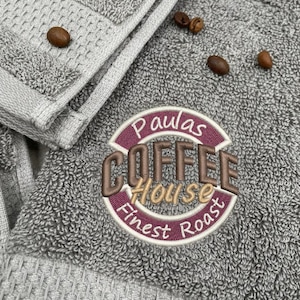 Könnte beinhalten: Ein graues, strukturiertes Handtuch mit einem gestickten kreisförmigen Logo, das in braunem, beigem und kastanienbraunem Garn Paulas COFFEE House Finest Roast zeigt. Mehrere geröstete Kaffeebohnen sind auf dem grauen Stoff verteilt.