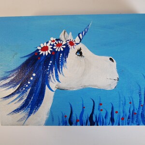 May include: A white unicorn with a blue mane and tail, wearing a flower crown with red and white flowers, is painted on a blue background with blue grass and red berries.