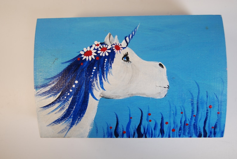 May include: A white unicorn with a blue mane and tail, wearing a flower crown with red and white flowers, is painted on a blue background with blue grass and red berries.