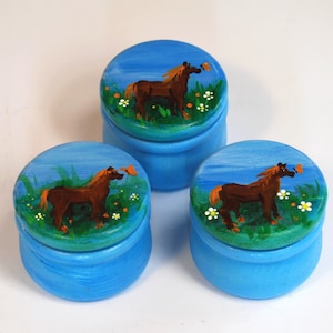 May include: Three small blue wooden boxes with lids. Each lid has a painted scene of a brown horse in a field with green grass and white flowers. A small orange butterfly is near each horse.