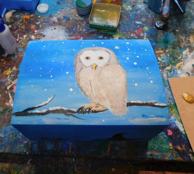 May include: A wooden box with a blue background and a white owl painted on the lid. The owl is perched on a branch with snow on it. The background is a blue sky with white snowflakes.