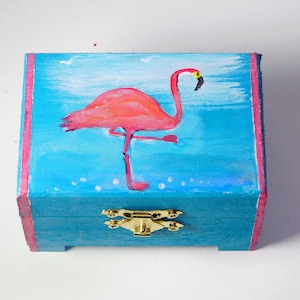 Könnte beinhalten: Eine kleine Holzkiste mit einem Scharnierdeckel und einem goldenen Verschluss. Die Kiste ist in einem leuchtenden Türkisblau lackiert, auf der Vorderseite ist ein rosa Flamingo gemalt. Der Flamingo hat einen gelben Schnabel und steht auf einem Bein.