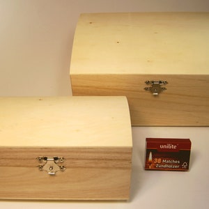 May include: Two wooden boxes with hinged lids and metal clasps. The smaller box is in front of the larger box. A small red box of matches is in the foreground.