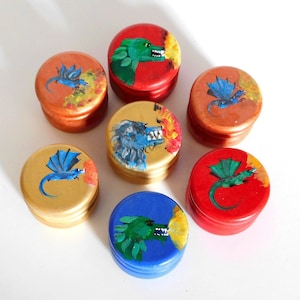 Puede incluir: Colección de siete pequeñas cajas redondas de madera con tapas. Cada caja está pintada de un color diferente y presenta un diseño de dragón pintado a mano. Los dragones están representados en varios colores, con llamas que emanan de sus bocas.