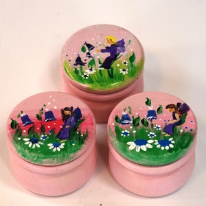 May include: Three pink wooden boxes with lids, each decorated with a different hand-painted fairy scene. The fairies are wearing purple dresses and have wings. The scenes include flowers, leaves, and bells.