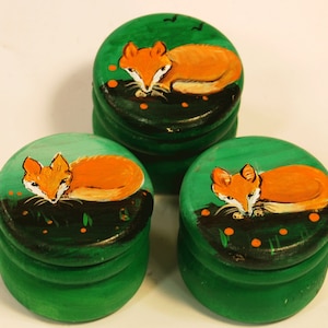 Op de afbeelding: Drie ronde houten dozen met groene deksels en vossenillustraties. Elke doos heeft een andere vos die op een groene achtergrond met oranje bloemen ligt.