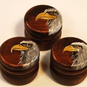 Op de afbeelding: Drie kleine houten doosjes met deksels, elk beschilderd met een ander profiel van een Amerikaanse zeearend. De adelaars hebben gele snavels en witte en grijze veren.
