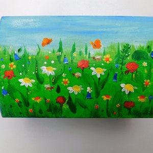 May include: A wooden box painted with a whimsical scene of a green meadow with colourful flowers, butterflies, and bees. The sky is blue with white clouds.