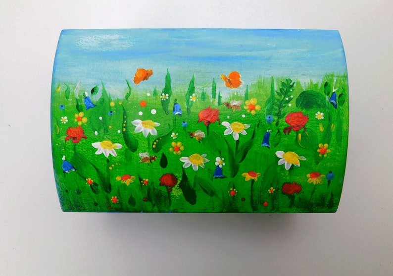 May include: A wooden box painted with a whimsical scene of a green meadow with colourful flowers, butterflies, and bees. The sky is blue with white clouds.