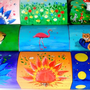 May include: Nine colourful wooden boxes with different painted designs. The boxes are stacked in three rows of three. The top row features a pink box with a blue flower, a green box with a field of flowers, and a green box with a grey cat and a red fox. The middle row features a green box with a lion, a blue box with a pink flamingo, and a blue box with an orange cat. The bottom row features a purple box with a blue flower, an orange box with a red sun, and a blue box with yellow circles.