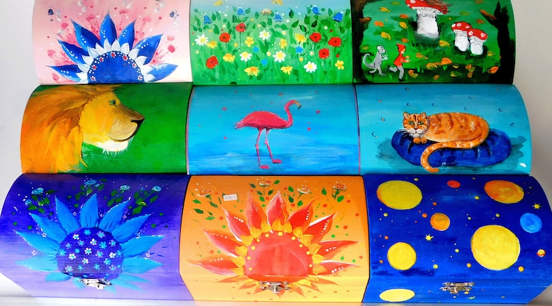 May include: Nine colourful wooden boxes with different painted designs. The boxes are stacked in three rows of three. The top row features a pink box with a blue flower, a green box with a field of flowers, and a green box with a grey cat and a red fox. The middle row features a green box with a lion, a blue box with a pink flamingo, and a blue box with an orange cat. The bottom row features a purple box with a blue flower, an orange box with a red sun, and a blue box with yellow circles.