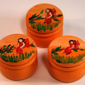 May include: Three small orange wooden boxes with lids. Each box is hand-painted with a fairy sitting on a branch with green leaves and red flowers.