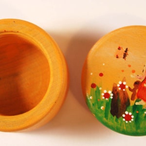 Op de afbeelding: Een houten doos met een beschilderd deksel. Het deksel heeft een fantasierijke scène met een fee met vleugels, een vlinder en bloemen in tinten groen, rood en wit.