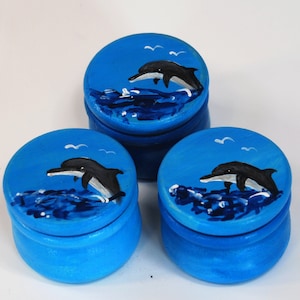 Op de afbeelding: Drie kleine blauwe houten doosjes met deksels, elk beschilderd met een zwart-witte dolfijn die uit het water springt. De doosjes hebben een witte achtergrond met blauwe golven en witte vogels.