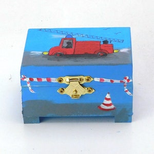 Könnte beinhalten: Eine kleine blaue Holzkiste mit einem roten Feuerwehrauto, das auf den Deckel gemalt ist. Die Kiste hat einen goldenen Verschluss und ein rot-weiß gestreiftes Band. Die Kiste steht auf einer weißen Oberfläche.