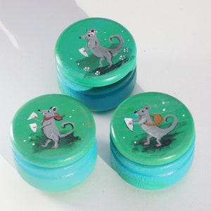 Op de afbeelding: Drie kleine ronde houten dozen met teal groene deksels en bodems. Elk deksel heeft een handgeschilderd ontwerp van een grijze muis die een brief vasthoudt en een rode sjaal draagt. De muizen bevinden zich op een groene achtergrond met witte stippen.