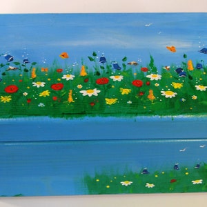 Könnte beinhalten: Eine blaue Holzkiste mit einem gemalten Bild eines Blumenfeldes. Die Blumen sind rot, gelb, weiß und blau. Auf der Kiste sind auch Schmetterlinge und Bienen gemalt.