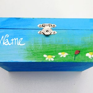 May include: A blue wooden box with a green and white painted scene of flowers and a ladybug. The box has a silver latch and the word "Name" is written on the front.