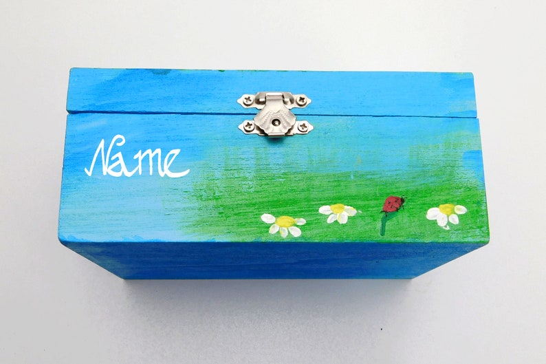May include: A blue wooden box with a green and white painted scene of flowers and a ladybug. The box has a silver latch and the word "Name" is written on the front.