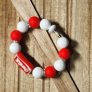 May include: A red and white beaded bracelet with a Coca-Cola charm. The bracelet has a silver clasp and is made of silicone beads.