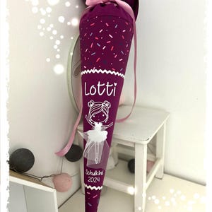 Könnte beinhalten: Lilafarbener Schultüte mit rosa und weißen Streuseln, einer weißen Ballerina-Illustration und dem Namen "Lotti" sowie dem Text "Schulkind 2024".