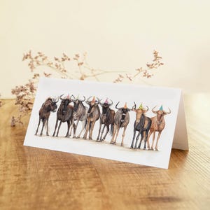May include: A folded greeting card featuring a watercolor illustration of several wildebeest wearing party hats. The animals are depicted in various shades of brown and black, standing in a row. The card is on a wooden surface.
