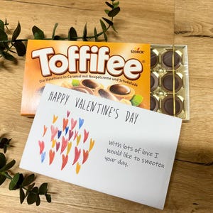 Pode incluir: Uma caixa de chocolates Toffifee com um cartão de Dia dos Namorados. O cartão tem ilustrações de corações coloridos e a mensagem "Happy Valentine's Day". Os chocolates estão numa bandeja dourada.