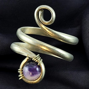 Könnte beinhalten: Ein silberfarbener Ring mit einem Spiraldesign. Der Ring hat eine gewickelte Fassung, die einen runden, lila Edelstein hält. Der Ring ist vor einem dunklen Hintergrund platziert.