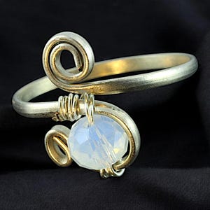 Op de afbeelding: Een zilveren draadgewikkelde armband met een witte opaal edelsteen. De armband heeft een spiraalvormig ontwerp en is gemaakt van draad.