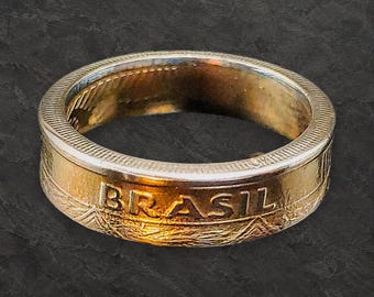 May include: A metallic ring crafted from a coin, featuring the word BRASIL embossed on the outer band. The ring has a polished gold-toned finish with a textured, ridged edge along the top and bottom rims, showcasing a distinct coin-like design.