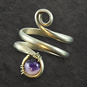 Könnte beinhalten: Ein silberfarbener Bypass-Ring mit einem Spiraldesign an einem Ende und einem runden, polierten violetten Amethyst-Edelstein, der am anderen Ende mit feinem Draht befestigt ist. Das Metallband hat eine glatte, reflektierende Oberfläche.