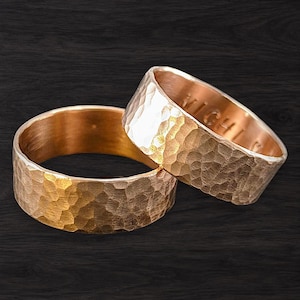 Könnte beinhalten: Zwei Ringe aus Roségold mit einer gehämmerten, texturierten Außenfläche. Die Ringe haben ein breites, flaches Banddesign. Die Innenseite des rechts positionierten Rings ist mit dem sichtbaren Text "MICHI" graviert. Das Metall hat einen warmen Glanz.