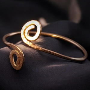 Può includere: Un bracciale dorato con una texture martellata e un design a spirale. Il bracciale è aperto e poggia su una superficie scura e strutturata. Il design a spirale è una caratteristica prominente.