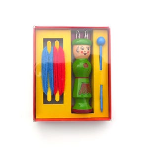 Puede incluir: Un juego de muñeca de punto de madera en una caja roja, amarilla y naranja. El juego incluye una muñeca verde con la cara pintada, un paquete de hilo azul y rojo y una herramienta de punto azul. La muñeca tiene un sombrero y un delantal verdes.