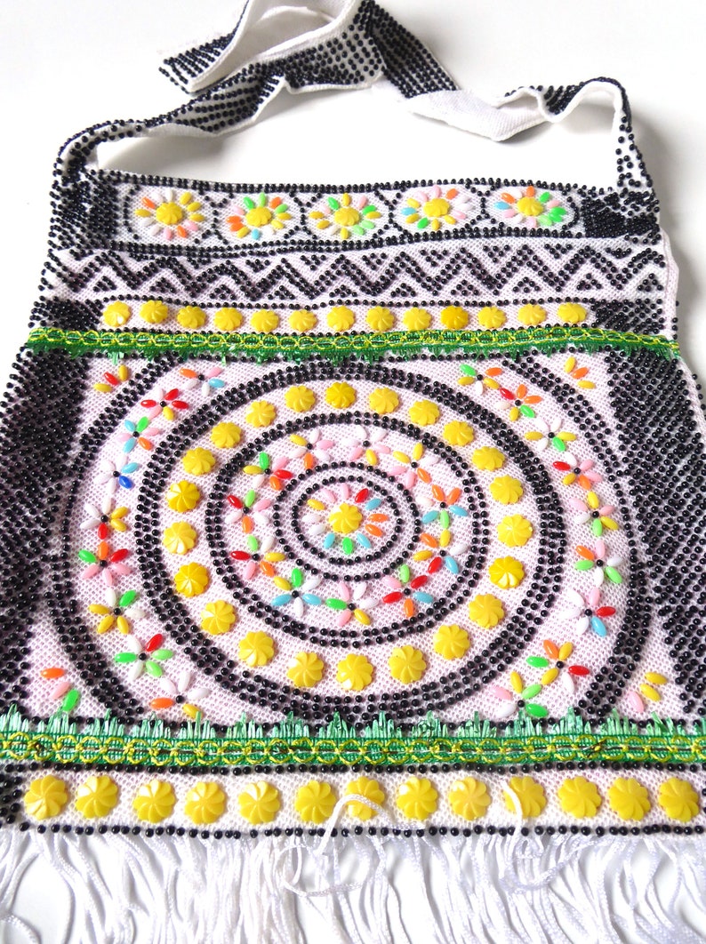 K&ouml;nnte beinhalten: Eine wei&szlig;e Perlenhandtasche mit einem schwarz-wei&szlig;en geometrischen Muster, einem gr&uuml;n-gelben Besatz und einem kreisf&ouml;rmigen Design aus gelben, rosa und mehrfarbigen Perlen.