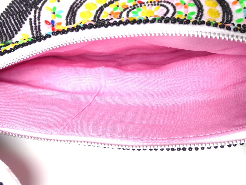 K&ouml;nnte beinhalten: Eine wei&szlig;e Perlenhandtasche mit einem rosa Stofffutter. Die Tasche hat einen Rei&szlig;verschluss und ist mit bunten Perlen verziert.