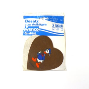 Könnte beinhalten: Eine Packung mit zwei braunen herzförmigen Bügelbildern mit einem Comicbild von Donald Duck. Die Packung ist blau und weiß und trägt die Aufschrift "Besatz zum Aufbugeln" und "wenco Qualität".