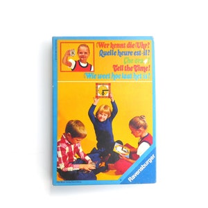 Könnte beinhalten: Eine farbenfrohe Spielschachtel mit blau-gelbem Design. Die Schachtel zeigt ein Bild von vier Kindern, die ein Spiel mit einer Uhr spielen. Der Text auf der Schachtel lautet "Wer kennt die Uhr? Quelle heure est-il? Che ora? Tell the Time! Wie weet hoe laat het is!" und "Ravensburger".