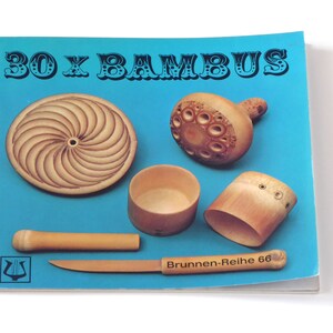 Puede incluir: Un libro azul con el t&iacute;tulo "30 x BAMBUS" en letras negras. La portada del libro presenta cinco tallas de madera diferentes hechas de bamb&uacute;, incluyendo un disco redondo, un taz&oacute;n peque&ntilde;o, un taz&oacute;n grande, un peque&ntilde;o objeto con forma de hongo y una pieza larga y delgada de bamb&uacute;.