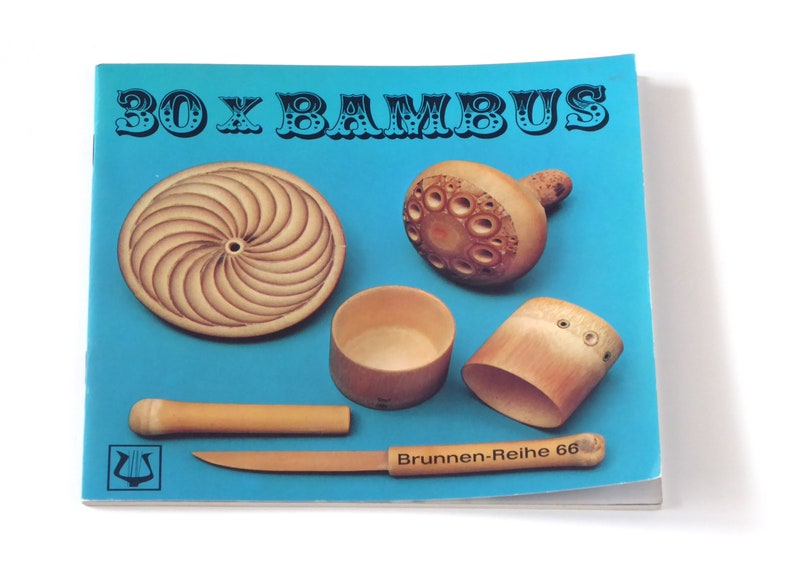 Puede incluir: Un libro azul con el t&iacute;tulo "30 x BAMBUS" en letras negras. La portada del libro presenta cinco tallas de madera diferentes hechas de bamb&uacute;, incluyendo un disco redondo, un taz&oacute;n peque&ntilde;o, un taz&oacute;n grande, un peque&ntilde;o objeto con forma de hongo y una pieza larga y delgada de bamb&uacute;.