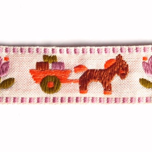 May include: A pink and white embroidered ribbon with a brown horse pulling a cart with orange boxes, and pink flowers on either side.