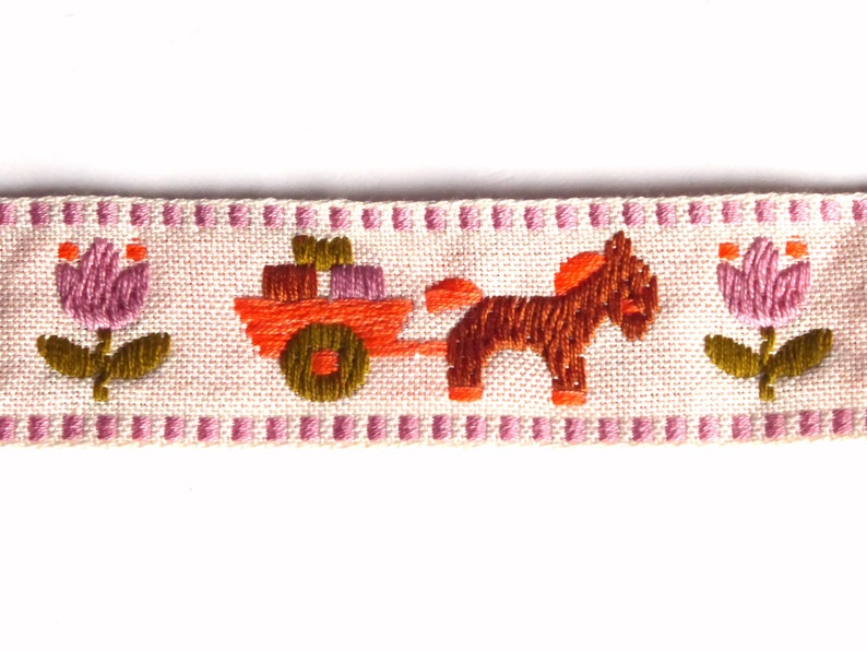 May include: A pink and white embroidered ribbon with a brown horse pulling a cart with orange boxes, and pink flowers on either side.